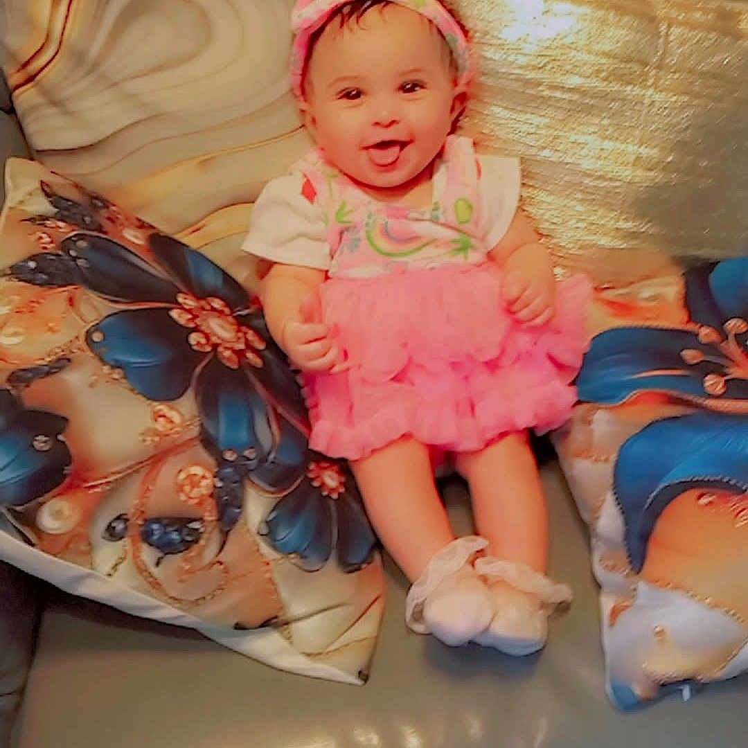 Beckyonce' is registered to the contest to win money with this photo: baby, child, colorful, couch, cute, floral_pattern, footwear, happy, headband, homedecor, indoors, infant, person, pillow, pink_dress, portrait, sitting, smiling, soft_toys, tongue_out