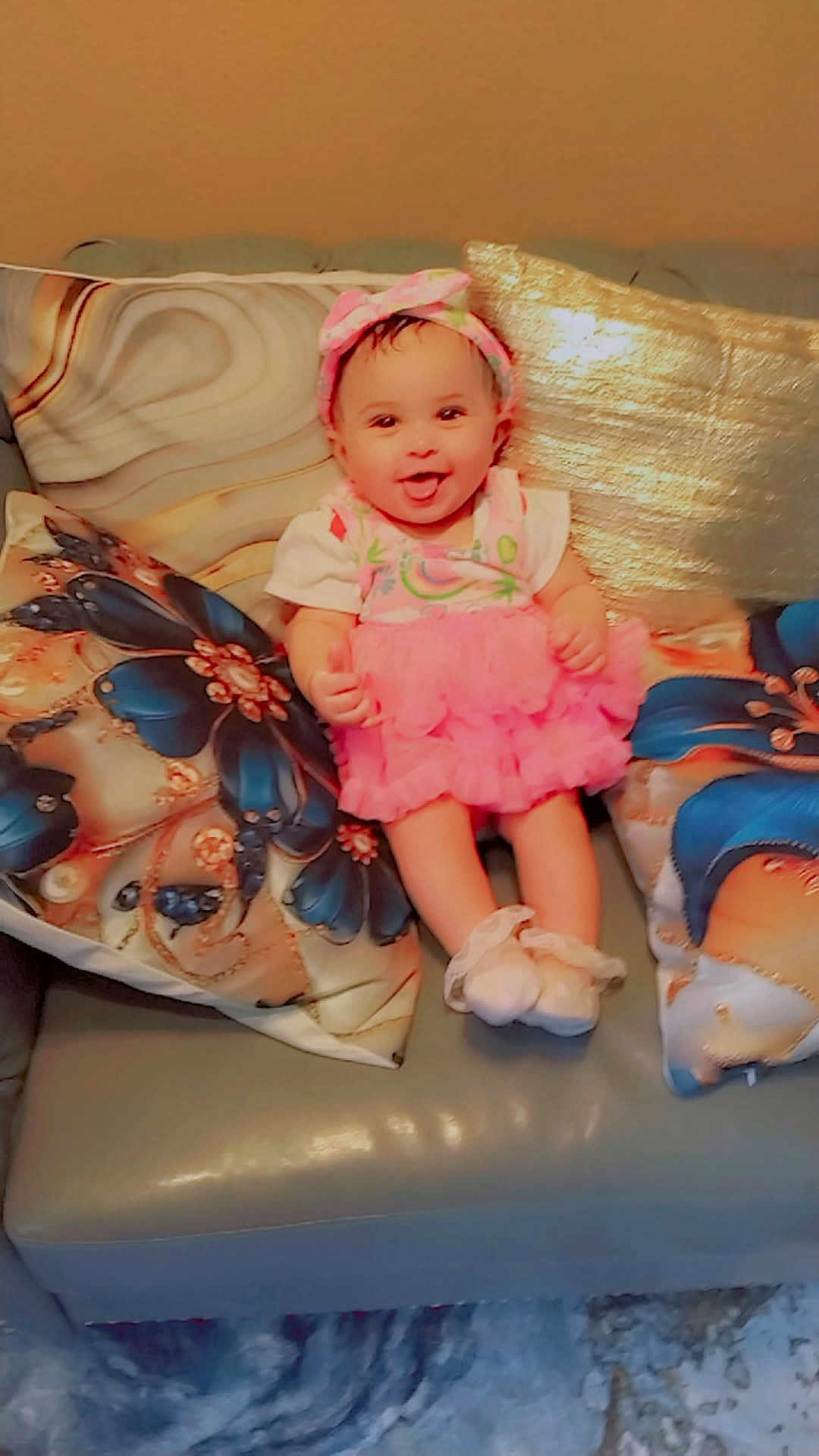 Beckyonce' is registered to the contest to win money with this photo: baby, child, pink_dress, headband, couch, pillow, floral_pattern, smiling, tongue_out, happy, indoors, sitting, cute, footwear, soft_toys, homedecor, colorful, infant, person, portrait