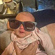 Braylen Farnsworth is registered to the contest to win money with this photo: baby, sunglasses, hand, indoor, mug, person, clothing, face, reflection, table, furniture, adult_hand, pink_clothing, newborn, closeup, casual, cozy, expression, holding, soft_lighting