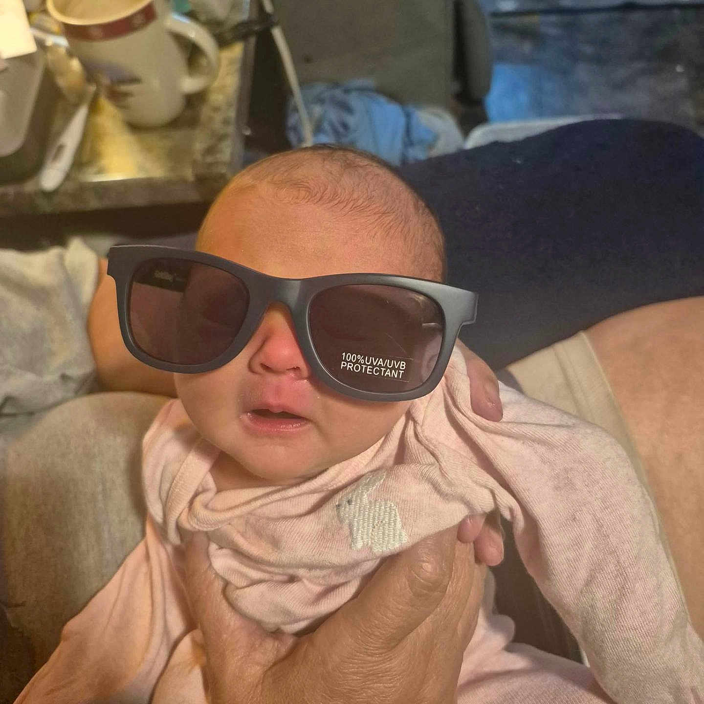 Braylen Farnsworth is registered to the contest to win money with this photo: baby, sunglasses, hand, indoor, mug, person, clothing, face, reflection, table, furniture, adult_hand, pink_clothing, newborn, closeup, casual, cozy, expression, holding, soft_lighting