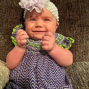 Nora is registered to the contest to win money with this photo: baby, smile, headband, flower, dress, purple, green, zigzag_pattern, hands, fists, couch, texture, infant, happy, portrait, sitting, cute, child, apparel, indoor