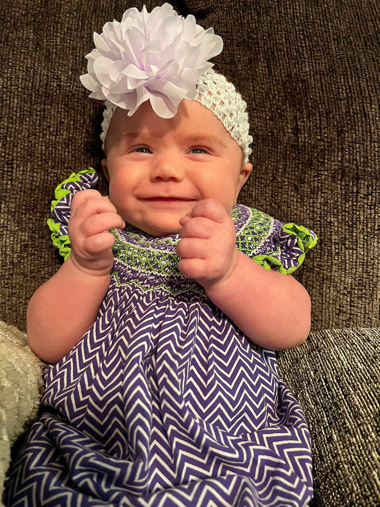 Nora is registered to the contest to win money with this photo: baby, smile, headband, flower, dress, purple, green, zigzag_pattern, hands, fists, couch, texture, infant, happy, portrait, sitting, cute, child, apparel, indoor