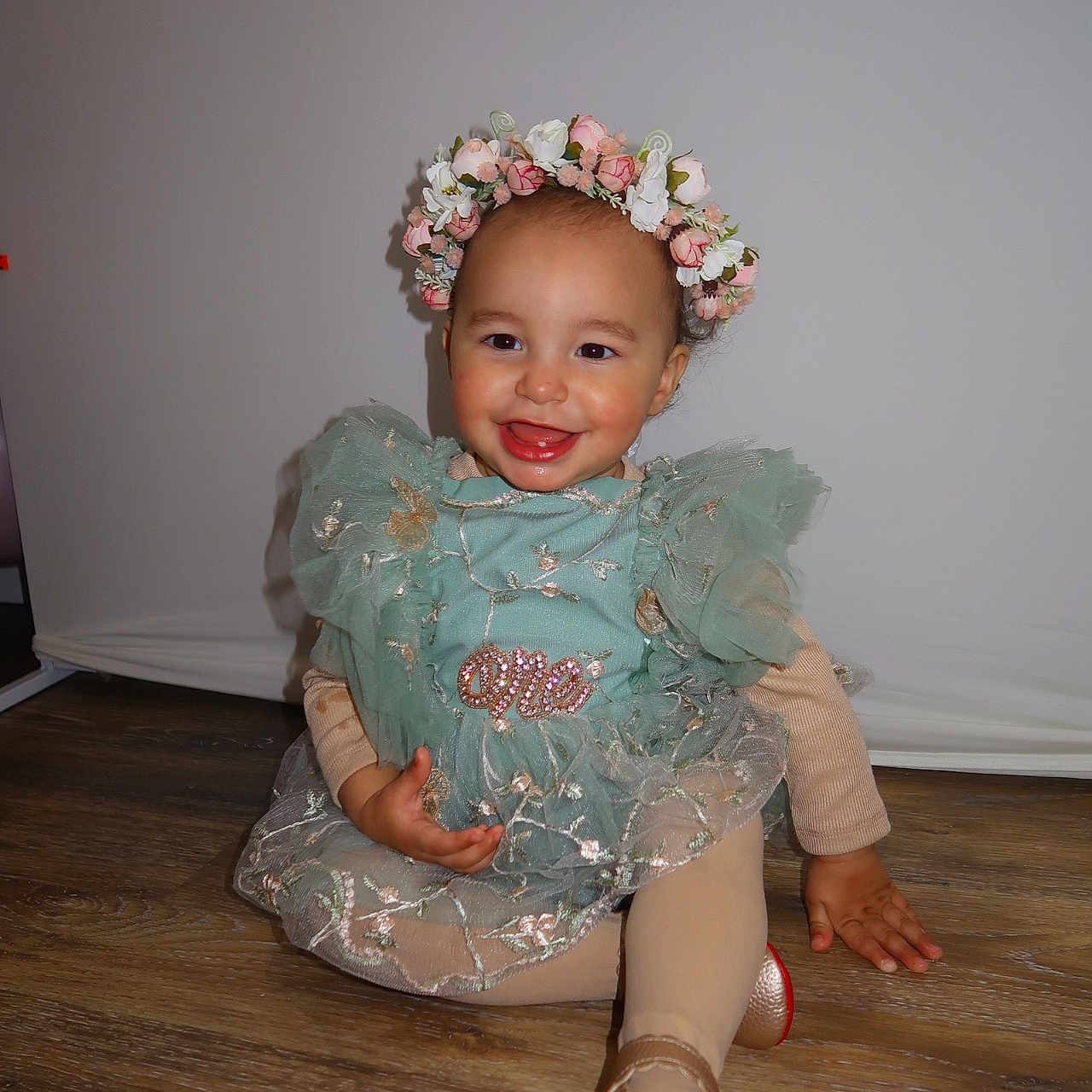 Francesca is registered to the contest to win money with this photo: toddler, child, smiling, dress, flower_crown, green_dress, sitting, wooden_floor, happy, portrait, cute, indoor, fashion, party, young_child, footwear, golden_shoes, long_sleeves, baby, playful