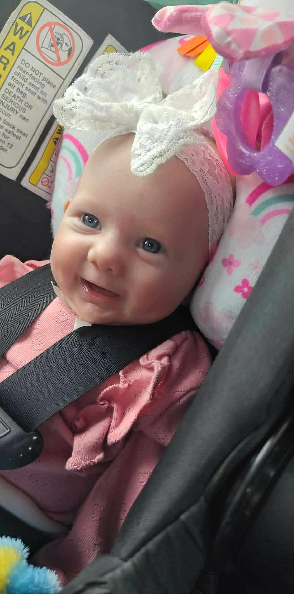 Maelee is registered to the contest to win money with this photo: accessory, baby, blue_eyes, bow, car_interior, car_seat, chubby_cheeks, closeup, headband, infant, lace_headband, pink_clothing, plush, portrait, rainbow_pattern, rosy_cheeks, safety_label, seatbelt, smiling, toy