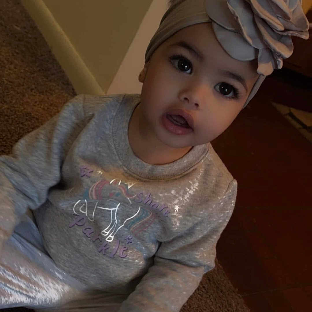 Cattleya Gómez is registered to the contest to win money with this photo: big_eyes, carpet, child, clothing, cozy, curious, cute, fashion, flower, headband, indoor, pants, person, portrait, sitting, sparkly, sweatshirt, toddler, unicorn, young_child