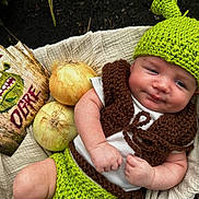 Everett is registered to the contest to win money with this photo: baby, infant, knitted_hat, green_hat, crocheted_vest, onions, wooden_sign, shrek, warning_sign, beige_blanket, outdoor, portrait, cute, baby_clothing, hand, face, eyes, smile, baby_boy, nature