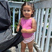 Sloane is registered to the contest to win money with this photo: toddler, child, outdoor, deck, wood, pink_clothing, shorts, sandals, grill_cover, railing, leaves, tree, summer, daylight, curly_hair, standing, person, portrait, expression, sunlight