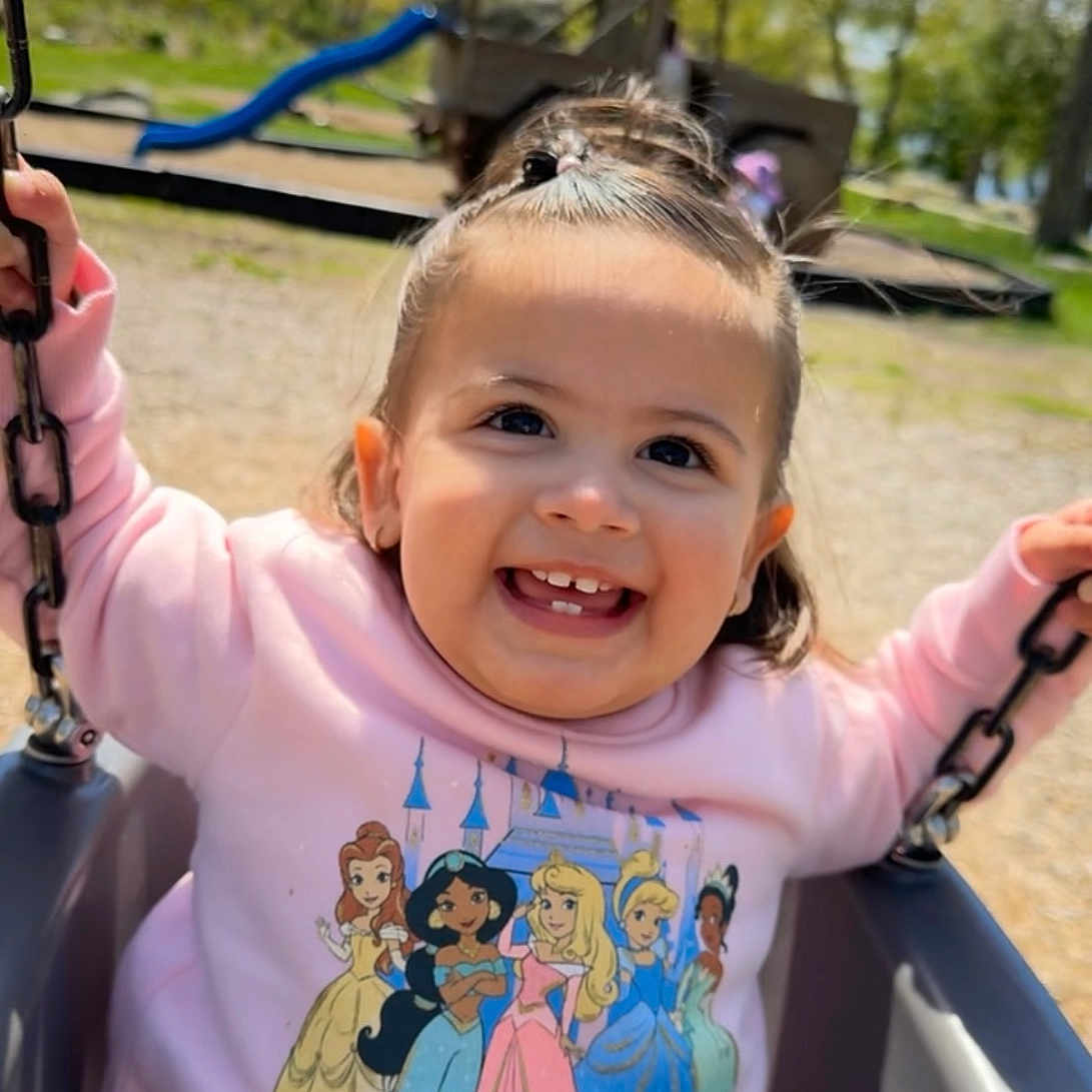Sloane is registered to the contest to win money with this photo: baby, casual_clothing, child, cute, daylight, disney_princesses, fun, happy, joy, outdoor, park, pink_sweatshirt, play_equipment, playground, smiling, sunny_day, swing, toddler, trees, young_child