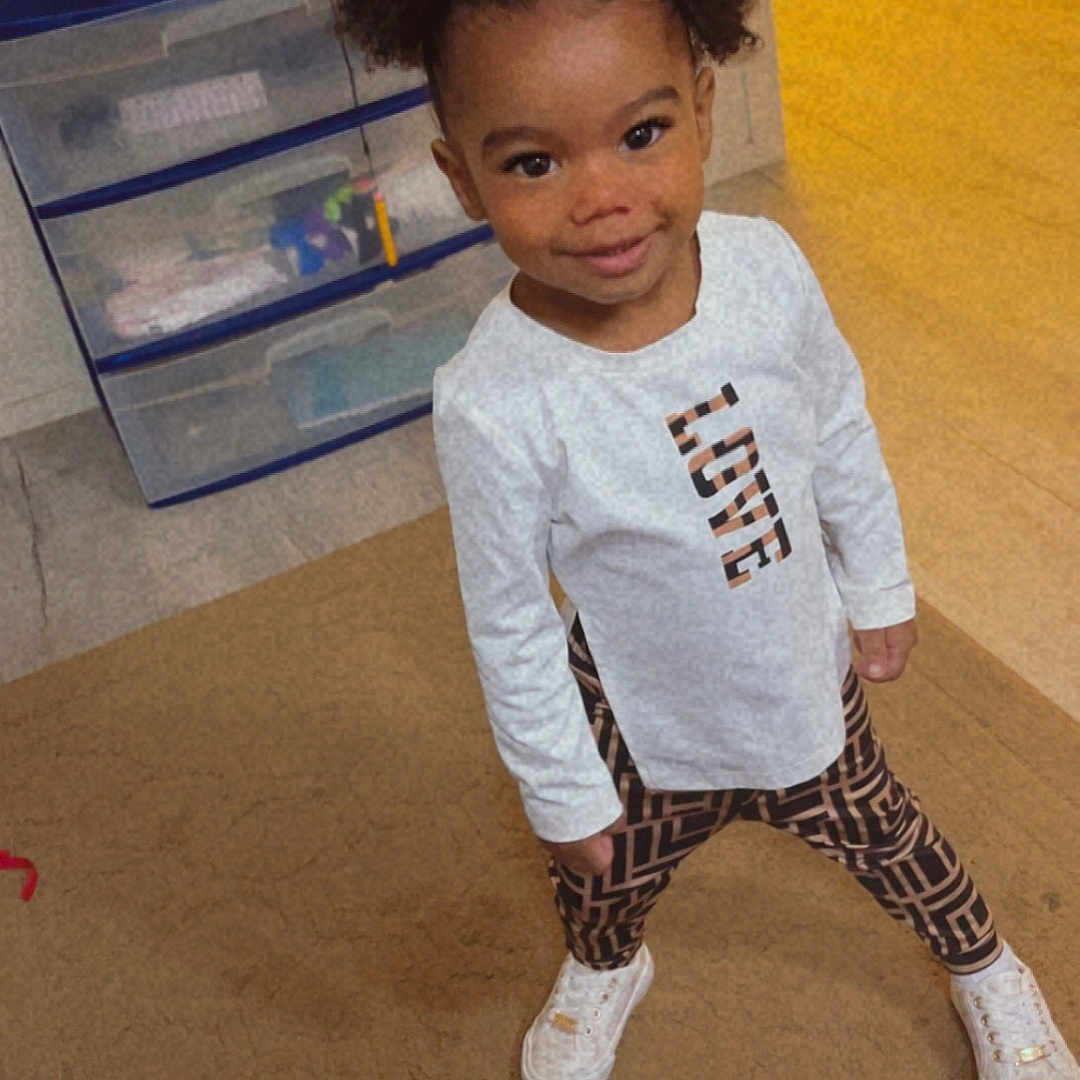 Omarianna is registered to the contest to win money with this photo: toddler, child, girl, pigtails, bows, white_shirt, patterned_leggings, sneakers, carpet, indoor, television, plastic_drawers, room, standing, smiling, cute, posing, floor, hand, nail_polish