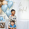 child, baby, balloons, heart_shaped_balloons, blue_balloons, beige_balloons, dinosaur_sweater, smiling_baby, holding, celebration, party_decoration, white_curtain, script_text, cute_kids, indoor, portrait, happy, siblings, playful, festive