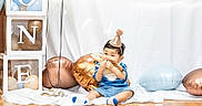 Simeon is registered to the contest to win money with this photo: baby, child, party_hat, balloon, blue_clothing, floor, wooden_floor, celebration, birthday, one_year_old, smiling, cute, indoor, white_backdrop, transparent_blocks, food, sitting, festive, sock, monkey_balloon