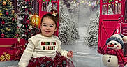 Vada is registered to the contest to win money with this photo: background, boots, child, christmas, decorations, festive, gift, green, holiday, indoor, portrait, red, skirt, smiling, snowman, sweater, toddler, tree, white, winter