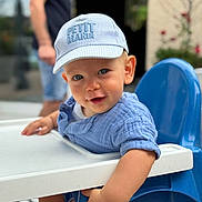 Sandro a rejoint le concours — aidez-le/la à gagner de superbes lots ! toddler, child, baby, blue_cap, highchair, smile, blue_eyes, short_sleeves, table, indoor, person, face, cute, curious, seated, furniture, adult_blurred, background, casual_clothing, portrait
