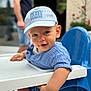 toddler, child, baby, blue_cap, highchair, smile, blue_eyes, short_sleeves, table, indoor, person, face, cute, curious, seated, furniture, adult_blurred, background, casual_clothing, portrait