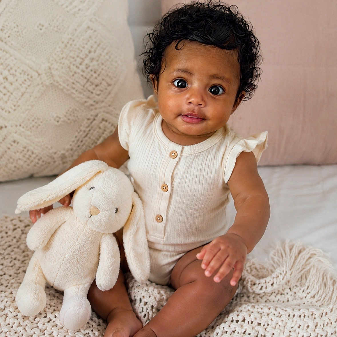 Amaris is registered to the contest to win money with this photo: baby, child, plush_bunny, knitted_blanket, pillows, curly_hair, sitting, indoors, cute, infant, neutral_colors, soft_toys, smiling, portrait, cozy, home, wooden_buttons, short_sleeves, barefoot, curious