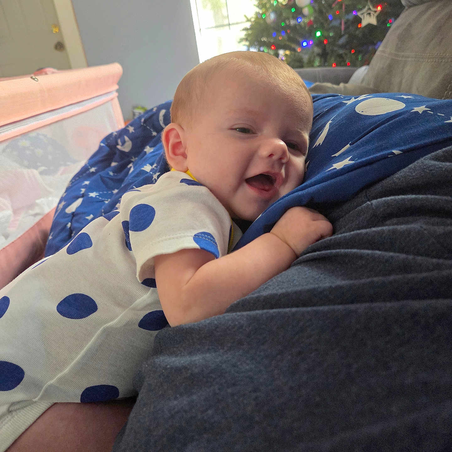 Mikailyn is registered to the contest to win money with this photo: arm, baby, carpet, christmas_tree, couch, cozy, daylight, decorations, face, family, hand, happy, indoor, infant, lights, onesie, person, polka_dot, smiling, window