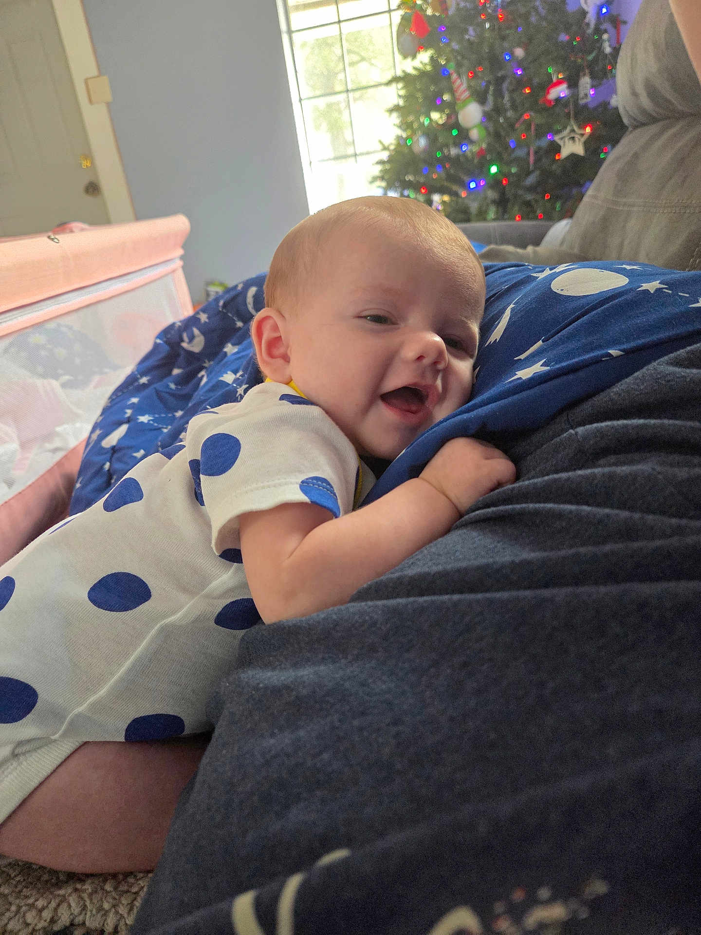 Mikailyn is registered to the contest to win money with this photo: baby, infant, polka_dot, onesie, smiling, face, arm, hand, indoor, christmas_tree, decorations, lights, couch, carpet, window, daylight, person, cozy, happy, family