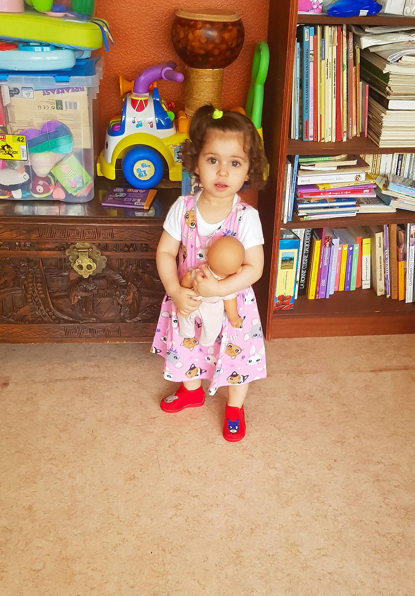 Neila participe au concours pour gagner de l'argent avec cette photo : baby_toddler_clothing, bookcase, child, clothing, dress, eye, floor, flooring, fun, head, leg, mammal, person, product, shelf, shelving, skin, standing, toddler, toy