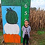 Emma joined the competition — help win amazing prizes! black_clothing, cardigan, child, clouds, cornfield, fall, farm, grass, green, height_measurement, nature, orange, outdoor, painting, pumpkin, shoes, sign, sky, smiling, white