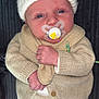 baby, infant, pacifier, knitwear, hat, pom_poms, beige, white, face, hand, clothing, cute, newborn, lying_down, portrait, child, soft_texture, warm_clothing, indoor, calm