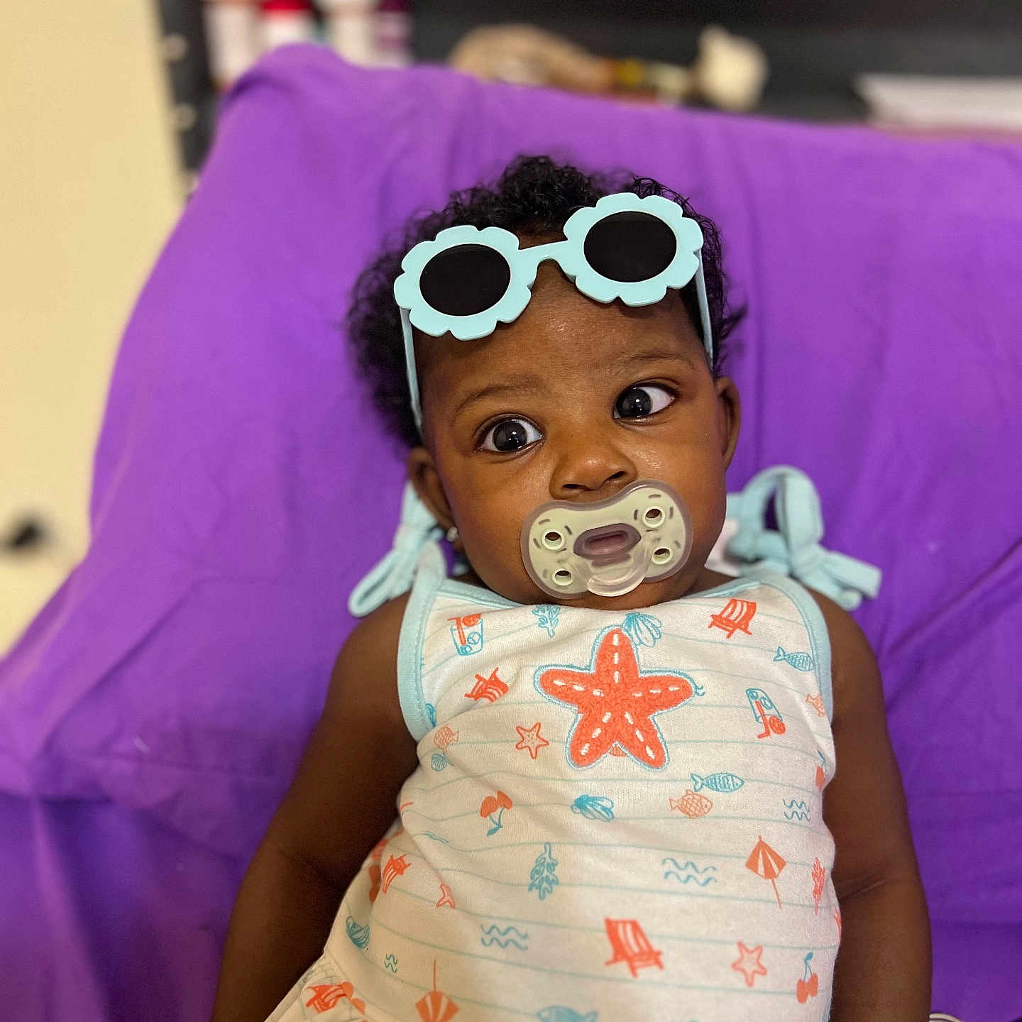 Aaleigha is registered to the contest to win money with this photo: accessory, baby, child, clothing, colorful, curly_hair, cute, expression, face, furniture, indoor, infant, pacifier, person, portrait, purple_chair, seated, starfish_outfit, sunglasses, toy