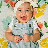 Delilah is registered to the contest to win money with this photo: baby, child, smiling, headband, bow, denim_overalls, clothing, happy, face, portrait, infant, cute, hands, colorful, blanket, floral_pattern, lying_down, indoors, person, young