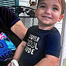 Asher is registered to the contest to win money with this photo: child, toddler, boy, smiling, portrait, outdoor, porch, seated, being_held, adult, t_shirt, text_on_clothing, toy, lantern, chair, house, siding, flowers, garden, happy