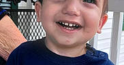 Asher is registered to the contest to win money with this photo: adult_hand, blue_shirt, brown_hair, child, closeup, daylight, face, family, happy, laughing, outdoor, porch, portrait, railing, shutters, smiling, super_cool_dude, teeth, text_on_clothing, toddler