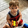 barefoot, black_shirt, blue_toy, boy, brown_hair, carpet, child, colorful, floor, indoors, looking_at_camera, playtime, portrait, red_clothing, serious_expression, shorts, sitting, toddler, toy, toy_truck