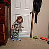 child, toddler, portrait, standing, smiling, hoodie, jacket, plaid, pants, sneakers, shoes, indoor, door, bedroom, carpet, toys, toy_car, stuffed_toy, dresser, clothing