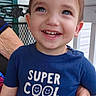 child, toddler, smiling, happy, portrait, blue_shirt, text_on_clothing, outdoor, porch, chair, adult_hands, family, brown_hair, blue_eyes, teeth, close_up, casual, summer, home, cute