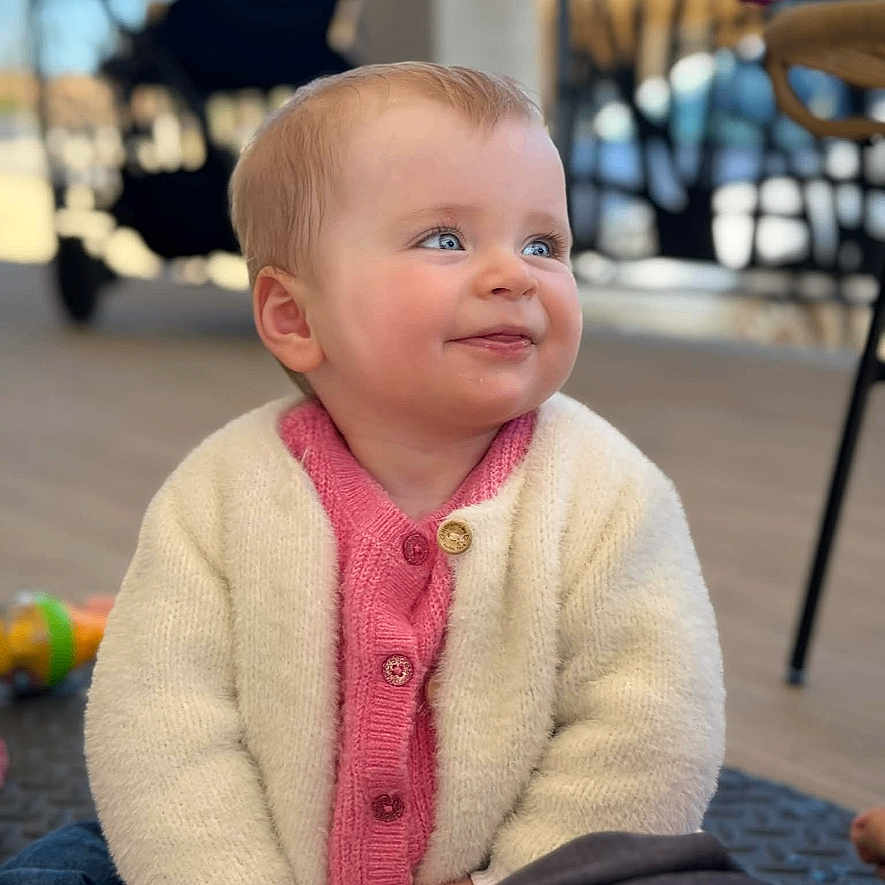 Lya a rejoint le concours — aidez-le/la à gagner de superbes lots ! baby, child, smiling, cardigan, pink, white, sitting, floor, indoor, portrait, happy, cute, person, blonde_hair, blue_eyes, toy, furniture, table, chair, stroller