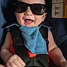 baby, child, sunglasses, car_seat, blue_bib, smiling, happy, infant, seatbelt, red_clothing, closeup, portrait, cute, indoor, safety_harness, cheerful, funny, young_child, accessory, casual