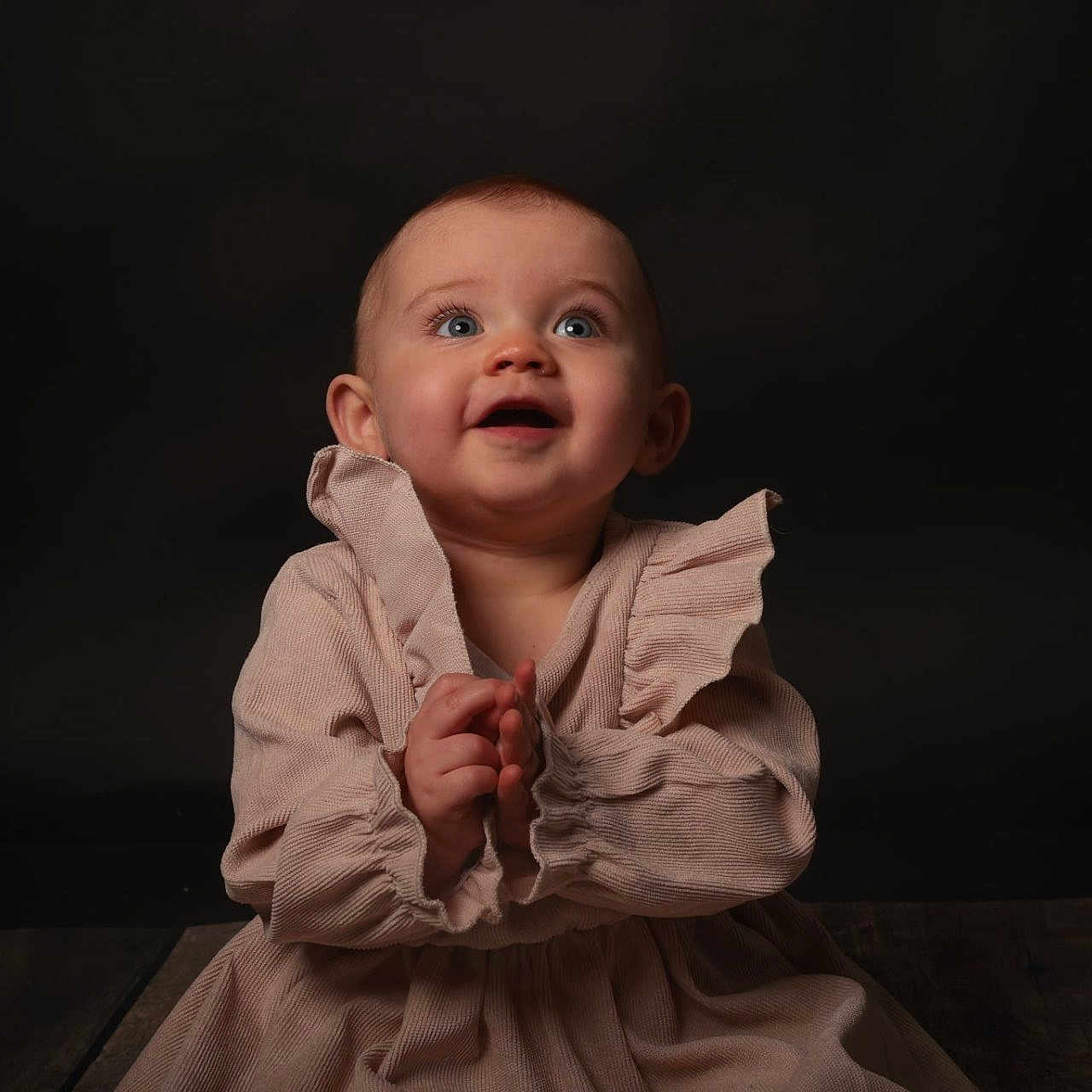 Thalia participe au concours pour gagner de l'argent avec cette photo : baby, background, blue_eyes, child, clapping, clothing, cute, dark, dress, face, hands, happy, human, infant, portrait, sitting, smiling, studio, wooden_floor, young