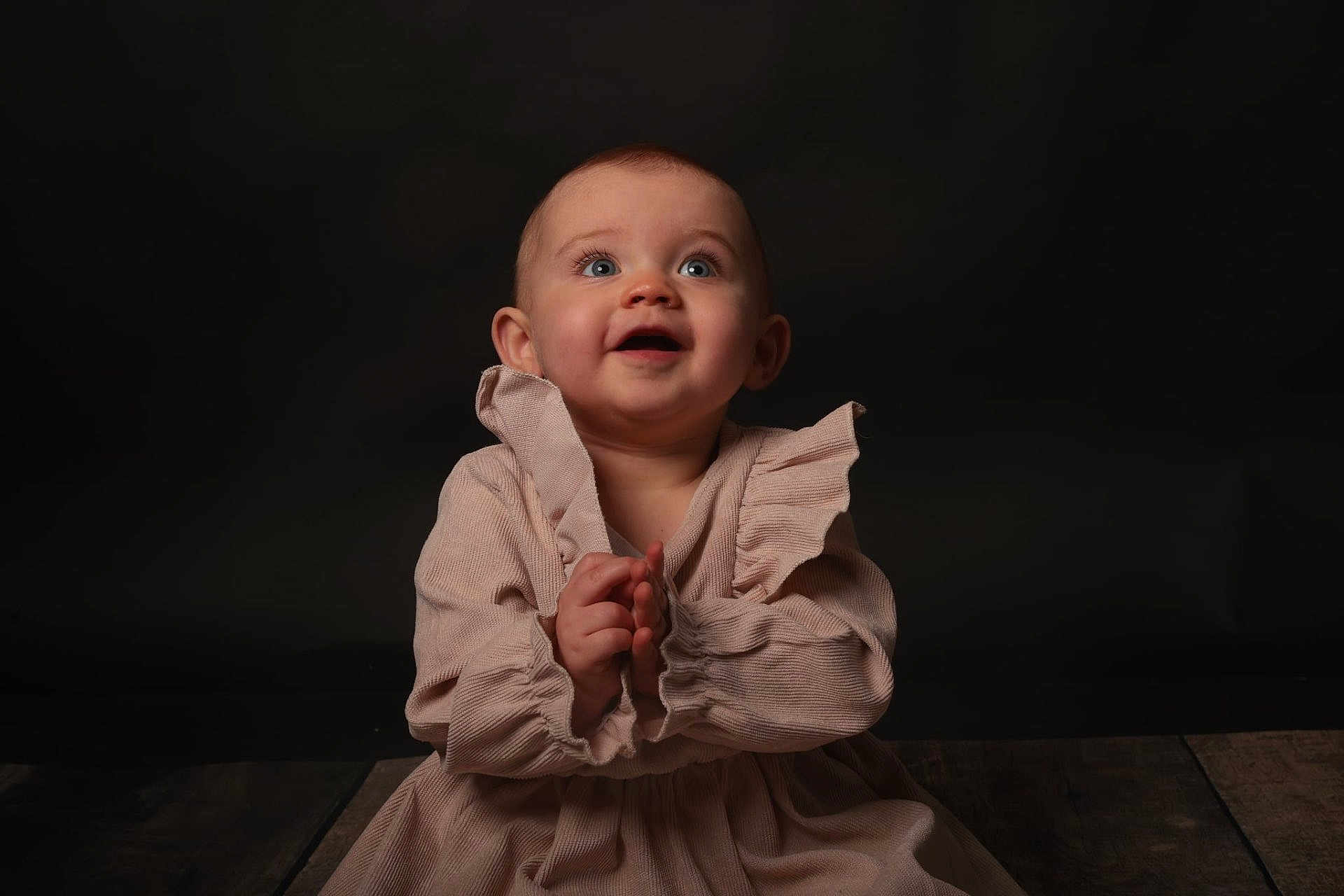 Thalia participe au concours pour gagner de l'argent avec cette photo : baby, child, clapping, dress, blue_eyes, happy, portrait, studio, wooden_floor, smiling, infant, cute, human, sitting, hands, face, young, clothing, background, dark