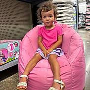 Kora joined the competition — help win amazing prizes! toddler, child, pink_bean_bag, curly_hair, pink_tshirt, shorts, white_sandals, store_aisle, product_boxes, floor, sitting, indoor, portrait, young_child, curious_expression, casual_clothing, retail, shopping, footwear, bean_bag_chair