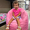 toddler, child, pink_bean_bag, curly_hair, pink_tshirt, shorts, white_sandals, store_aisle, product_boxes, floor, sitting, indoor, portrait, young_child, curious_expression, casual_clothing, retail, shopping, footwear, bean_bag_chair