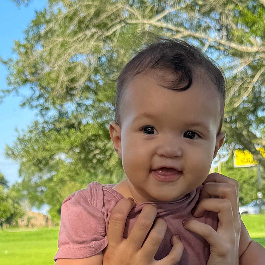 Kora is registered to the contest to win money with this photo: baby, child, person, smile, happy, outdoor, park, green_grass, tree, blue_sky, sunlight, holding, shoulders, casual_clothing, nature, daytime, cute, portrait, head, human_hand