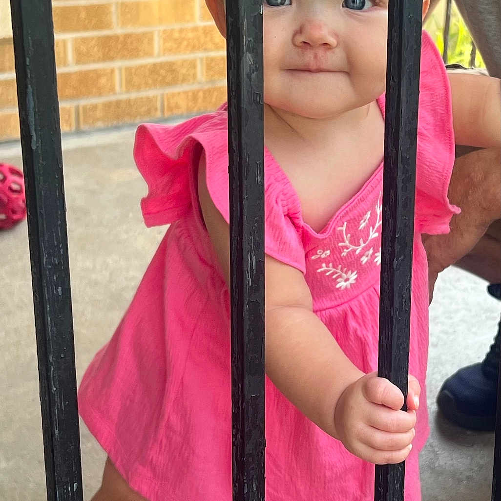 Lakelynne is registered to the contest to win money with this photo: baby, blue_eyes, brick_wall, child, curious, cute, daylight, footwear, hand, holding, metal_bars, outdoor, person, pink_dress, portrait, sandals, skin, standing, summer, toddler