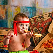 James is registered to the contest to win money with this photo: baby, child, pizza, pepperoni, food, eating, headband, red, green, cardboard_box, background, colorful, happy, smiling, indoor, person, clothing, sitting, cheese_pizza, hands