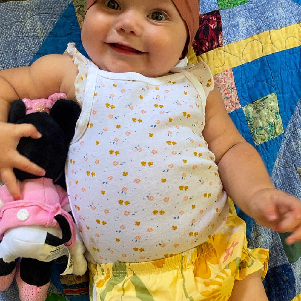 Kaylonni joined the competition — help win amazing prizes! baby, bow, child, clothing, colorful, cute, floral_pattern, happy, headband, indoors, infant, minnie_mouse, patchwork, person, plush_toy, quilt, smiling, toddler, white_tank_top, yellow_shorts