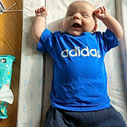Marshall is registered to the contest to win money with this photo: baby, infant, blue_shirt, adidas, smiling, arms_up, changing_pad, wooden_floor, baby_wipes, happy, cute, laying_down, patterned_pad, clothing, child, person, indoor, bright, young, cheerful