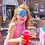 child, girl, face_paint, pink_dress, red_drink, straw, sandwich, outdoor, sunny, community_centre, table, chair, curly_hair, smiling, food, drink, pink_bag, daylight, happy, person