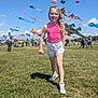 child, girl, smiling, outdoor, grass, field, kite, colorful_kites, festival, people, sunny, blue_sky, shorts, tank_top, sneakers, walking, happy, event, daytime, crowd