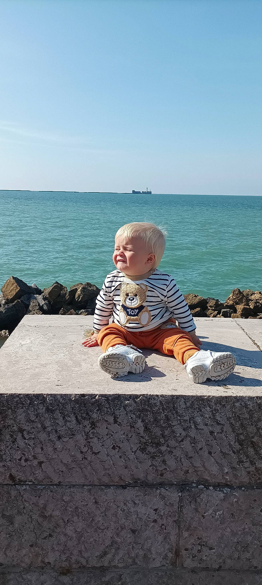 Timothée a rejoint le concours — aidez-le/la à gagner de superbes lots ! asphalt, baby, baby_toddler_clothing, child, coast, flooring, fun, happy, horizon, leisure, ocean, person, sitting, sky, soil, summer, t_shirt, toddler, travel, water