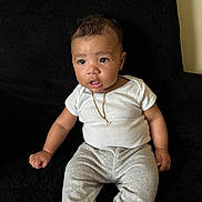 Legend is registered to the contest to win money with this photo: baby, child, infant, sitting, white_shirt, gray_pants, gold_necklace, letter_l_pendant, black_chair, curious_expression, indoors, cute, portrait, person, furniture, young_child, casual_clothing, comfortable, skin, hair