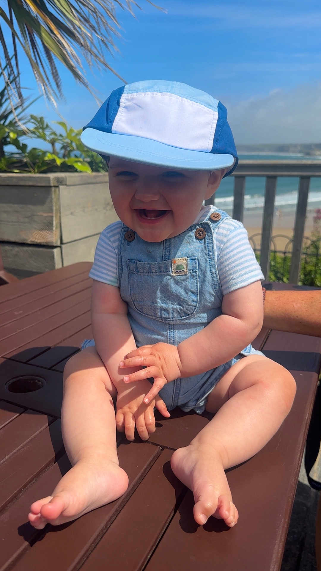 Arlo joined the competition — help win amazing prizes! baby, child, smiling, cap, denim_overalls, outdoor, table, wood, sunlight, beach, sky, plants, hand, foot, person, summer, happy, nature, portrait, seaside