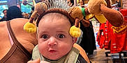 Anthony is registered to the contest to win money with this photo: baby, infant, child, adult_hand, headband, stuffed_animal, face, eyes, bib, surprised_expression, indoor, store, retail, brick_wall, wood_floor, window, lights, clothing, person, portrait