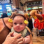 baby, infant, child, adult_hand, headband, stuffed_animal, face, eyes, bib, surprised_expression, indoor, store, retail, brick_wall, wood_floor, window, lights, clothing, person, portrait
