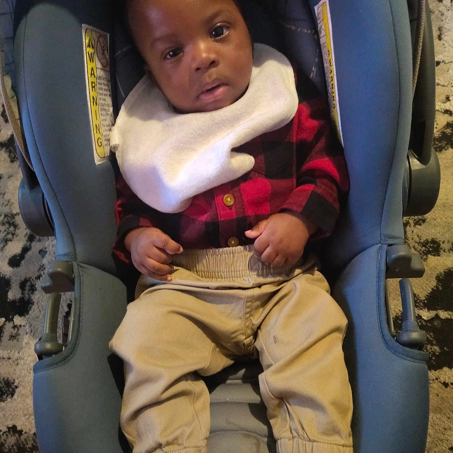 Kingston S Hicks is registered to the contest to win money with this photo: baby, bib, car_seat, carpet, checkered_shirt, child, cozy, dark_hair, eyes, face, floor, hands, indoor, infant, khaki_pants, looking_at_camera, plaid_shirt, portrait, socks, toddler